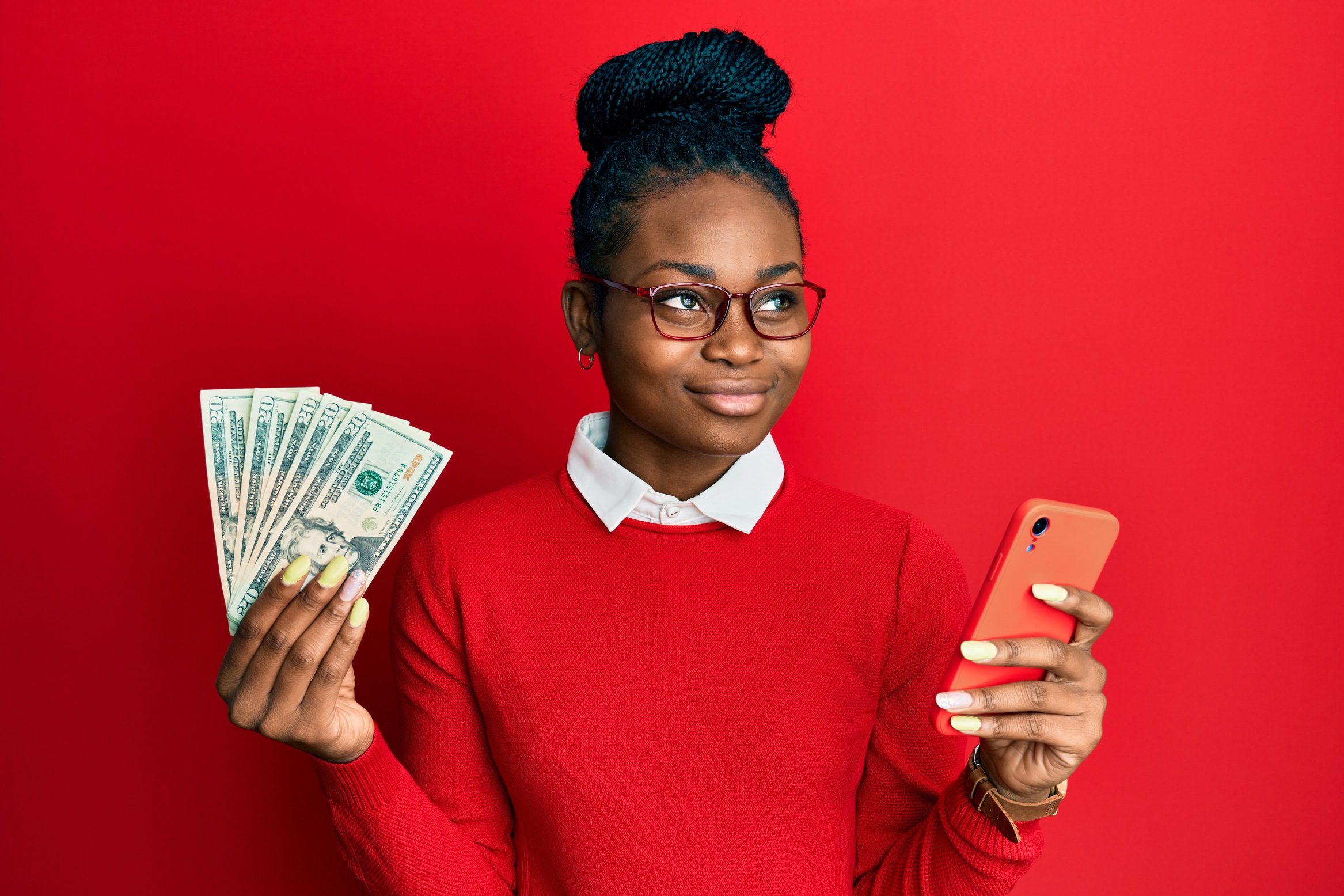 Young african american woman using smartphone holding usa dollars smiling looking to the side and staring away thinking.